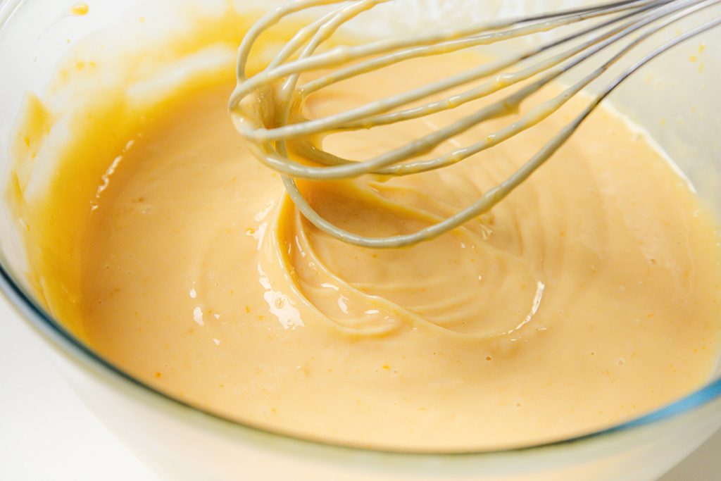A metal whisk stirs a thick, creamy, light orange batter in a clear glass bowl&mdash;perfect for making an easy lemon pie.