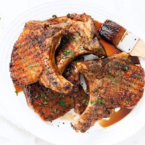 Grilled Pork Chops arranged on a white plate, garnished with chopped parsley. A basting brush and a cup of Sriracha Soy Glaze sit beside the chops, creating a bright, fresh scene with bold, appetizing flavors.