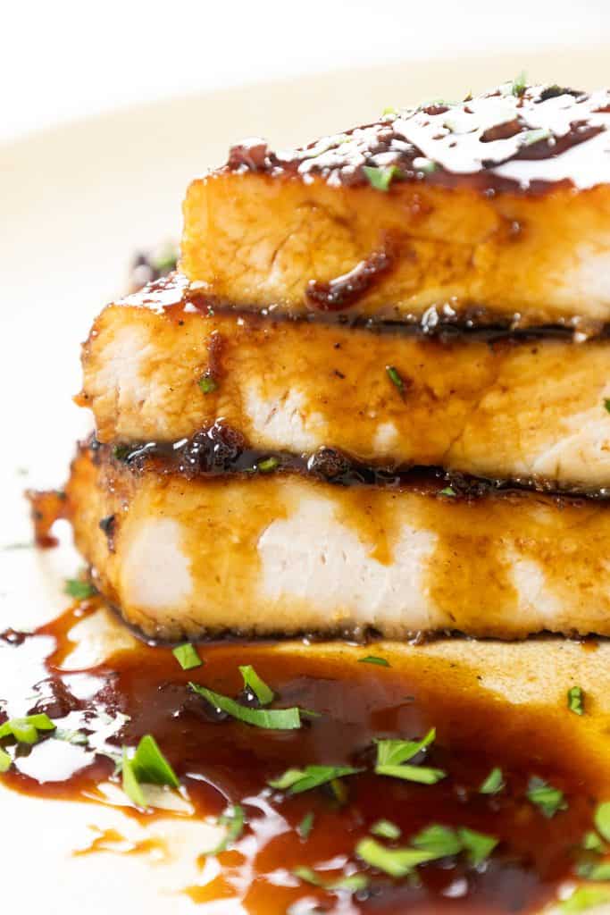 Three thick slices of cooked chicken breast stacked on a plate, topped with a glossy Sriracha Soy Glaze and garnished with finely chopped herbs.