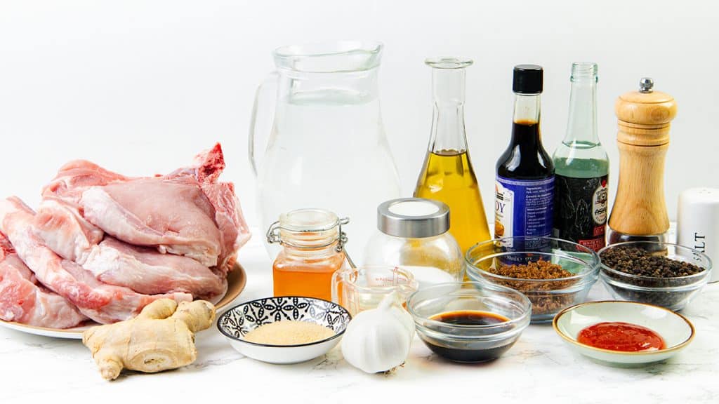 A variety of ingredients on a white surface, including raw brined pork chops, ginger, garlic, sauces, vinegar, soy sauce, honey, brown sugar, water, ground pepper, salt, and a small bowl of Sriracha soy glaze.
