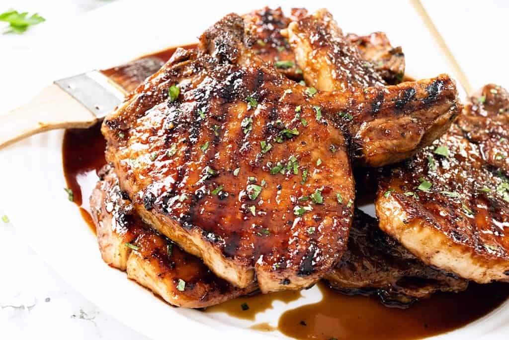 Brined pork chops are grilled to perfection and glazed with a shiny Sriracha Soy Glaze, then stacked on a white plate and garnished with chopped herbs. A basting brush, coated in the flavorful sauce, rests nearby.