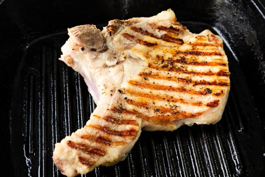 A grilled pork chop with seared grill marks is cooking on a black ridged grill pan. The brined pork chop is lightly seasoned, features a golden brown crust, and promises extra juiciness and flavor in every bite.
