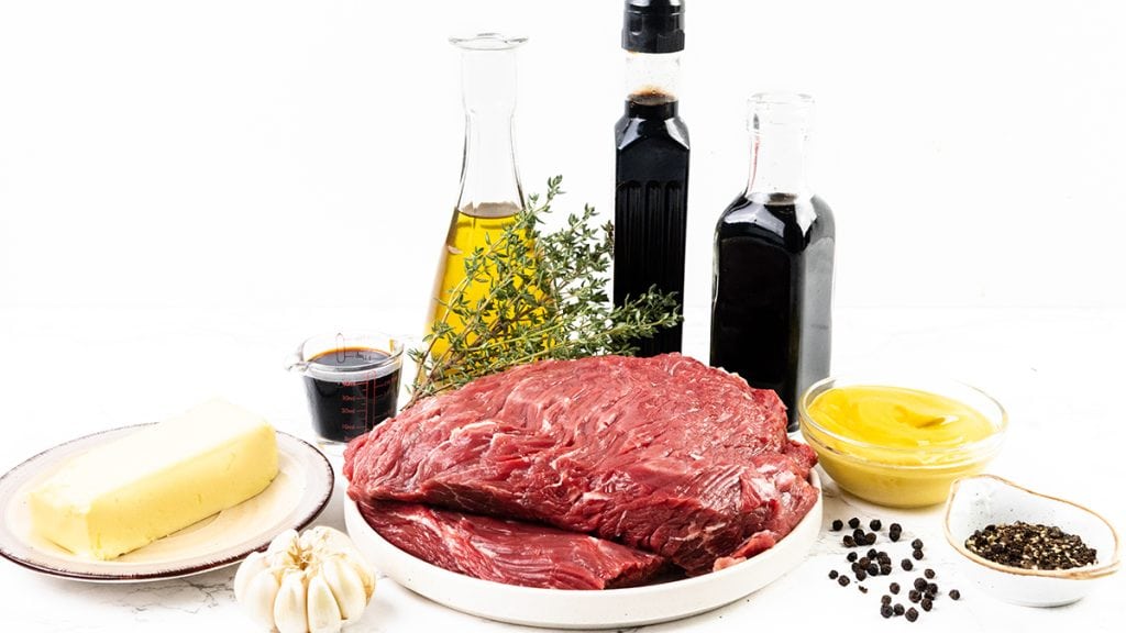 Two raw steaks on a plate surrounded by butter, garlic, fresh thyme, olive oil, balsamic vinegar, soy sauce, Dijon mustard, and black peppercorns&mdash;perfect for making flavorful Skillet Marinated Steak on a white background.