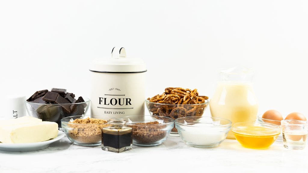 A variety of baking ingredients on a white surface, including flour, butter, chocolate, pretzels, brown and white sugar, eggs, milk, vanilla extract, melted butter, and salt—perfect for making salted caramel or chocolate pretzel bars.