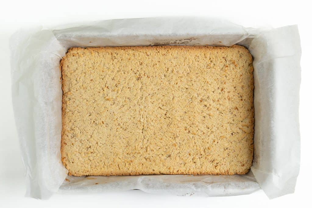A rectangular, golden-brown Salted Caramel oat bar sits in a baking pan lined with parchment paper, viewed from above on a white background.