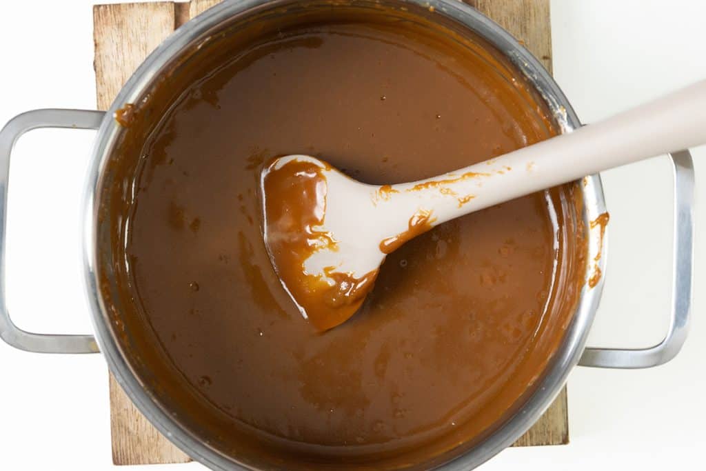 A pot filled with smooth Salted caramel sauce being stirred with a white spatula, perfect for drizzling over chocolate pretzel bars, sits on a wooden board.