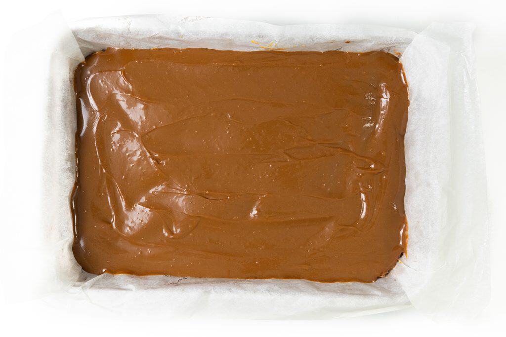 A rectangular baking pan lined with parchment paper, filled with a smooth layer of salted caramel mixture, ready to be set or baked into decadent salted caramel bars.