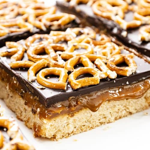 A close-up of Chocolate Pretzel Bars featuring a cookie base, a layer of salted caramel, glossy chocolate, and pretzel twists on top. The bars are cut into squares and arranged closely together.
