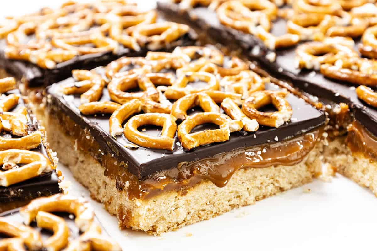 A close-up of Chocolate Pretzel Bars featuring a cookie base, a layer of salted caramel, glossy chocolate, and pretzel twists on top. The bars are cut into squares and arranged closely together.
