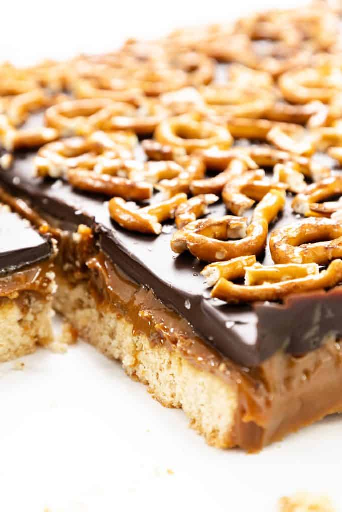A close-up of chocolate pretzel bars with a shortbread base, gooey salted caramel layer, thick chocolate topping, and mini pretzels. One piece has been cut and partially removed from the bar.