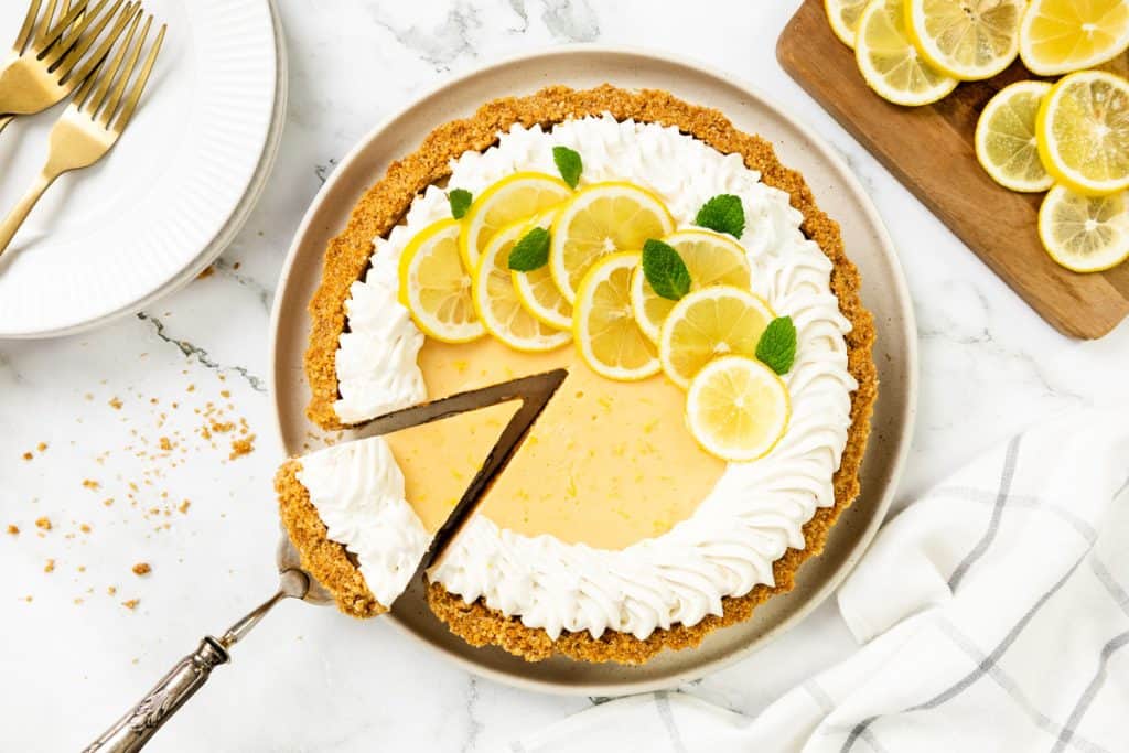 An Easy Lemon Pie with a graham cracker crust, topped with whipped cream, lemon slices, and mint leaves. One slice is served on a pie server, with plates, forks, and extra lemon slices nearby on a marble surface.