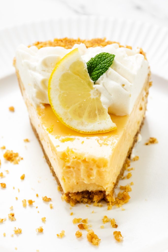 A slice of Easy Lemon Pie with a graham cracker crust, topped with whipped cream, a lemon wedge, and a mint leaf, served on a white plate. Crumbs are scattered around the slice.