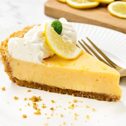 A slice of creamy Easy Lemon Pie with a graham cracker crust, topped with whipped cream, a lemon slice, and a mint leaf, sits on a white plate with a gold fork; extra lemon slices are in the background.