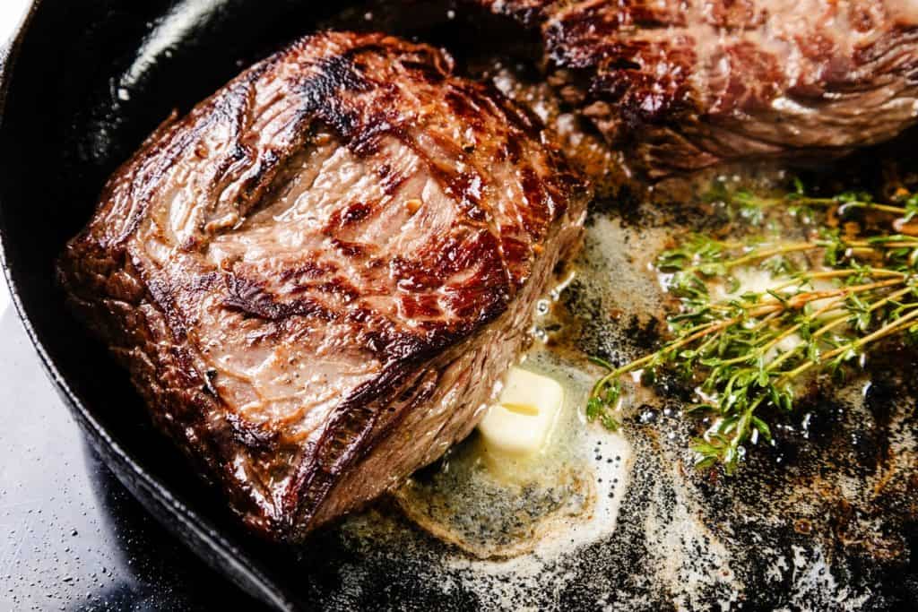A seared skillet steak cooks in a cast iron pan with melted butter, sprigs of fresh thyme, and a clove of garlic&mdash;an easy steak recipe for rich, savory flavor.