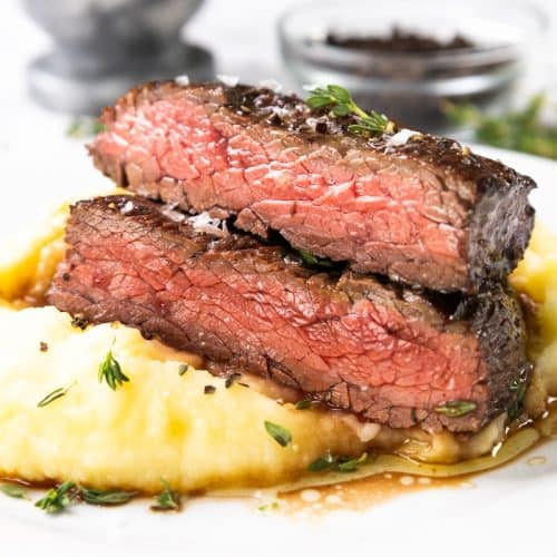 Two thick slices of marinated steak rest on a bed of creamy mashed potatoes, garnished with fresh thyme and drizzled with sauce on a white plate, bringing skillet-cooked perfection to every bite.
