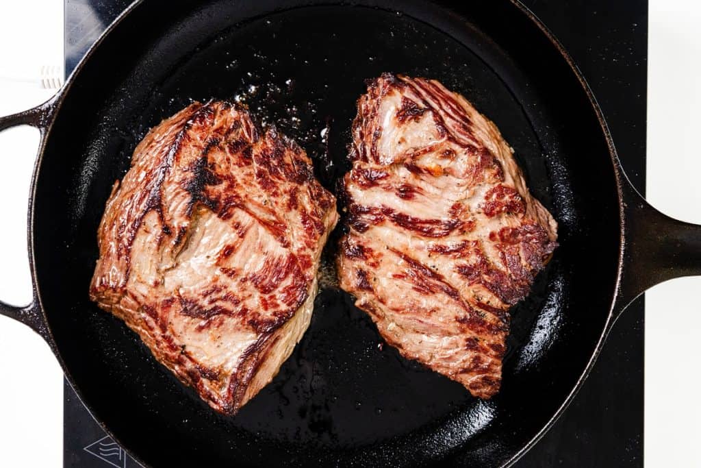 Two marinated steaks with char marks sizzle in a black cast iron skillet, viewed from above as they cook to perfection on the stovetop.