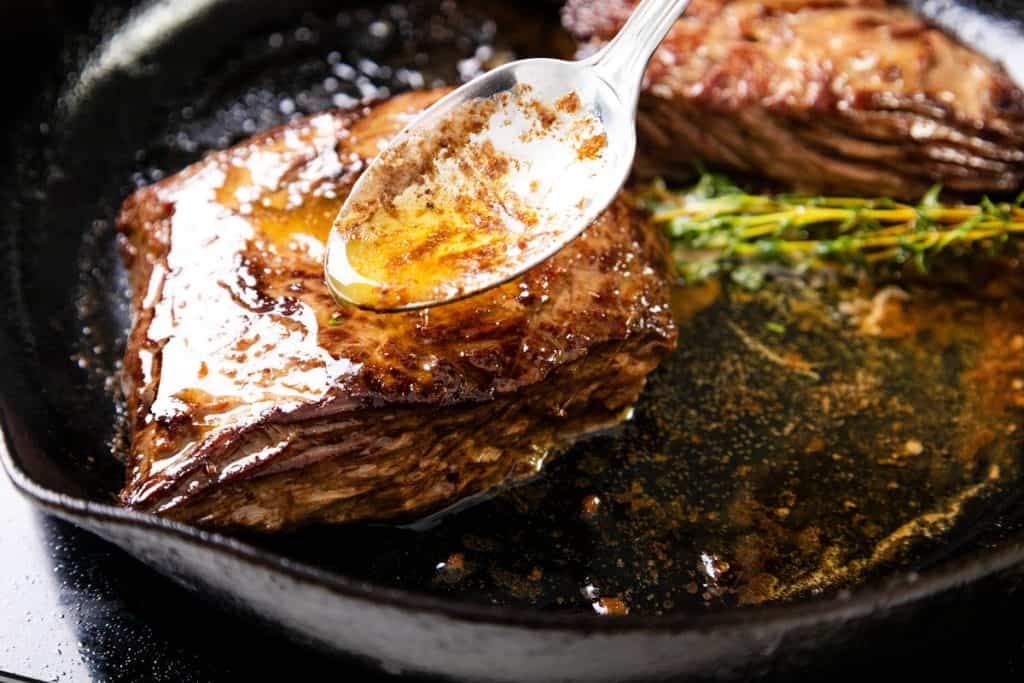 A spoon drizzles melted butter and seasonings over a marinated steak in a cast-iron skillet, with fresh thyme visible beside the meat.