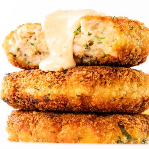 Three golden-brown, breaded salmon cakes are stacked on a white plate. The top cake is cut in half, revealing a moist interior, and is topped with a dollop of spicy aioli.