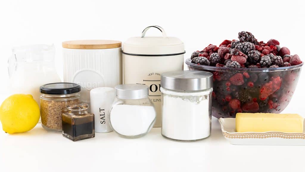 Assorted baking ingredients for berry cobbler, including a bowl of mixed berries, butter, jars, and containers with sugar, flour, salt, baking powder, a lemon, vanilla extract, brown sugar, and milk, arranged on a white surface.