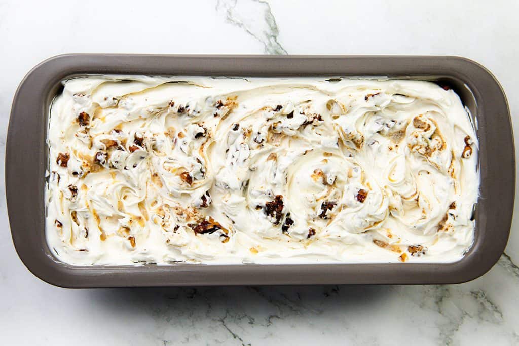 A gray rectangular pan filled with swirled, creamy white Butter Pecan Ice Cream mixed with visible chunks of cookies or nuts sits on a marble surface.