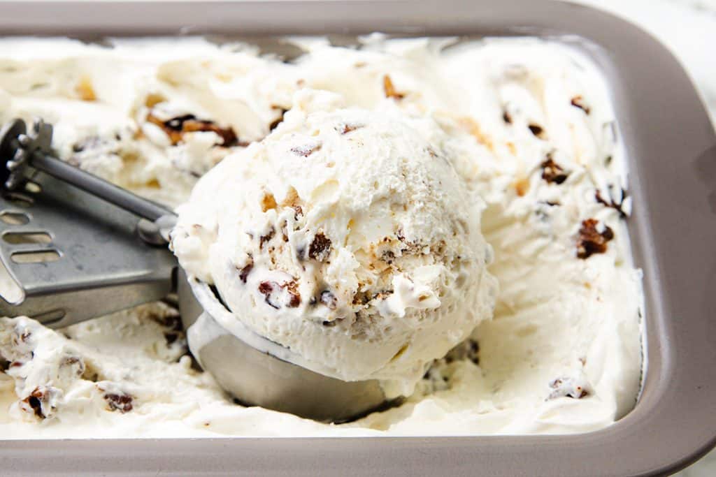 A metal scoop holds a large serving of creamy Butter Pecan ice cream with chocolate chunks and nuts, resting in a tray filled with more rich and indulgent ice cream.