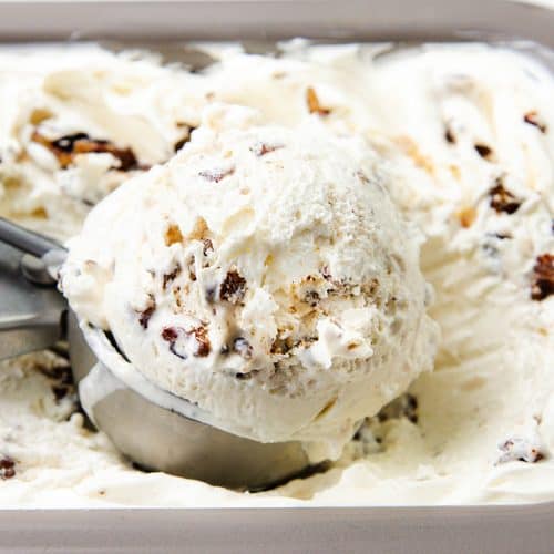 A metal scoop holds a large serving of creamy Butter Pecan ice cream with chocolate chunks and nuts, resting in a tray filled with more rich and indulgent ice cream.