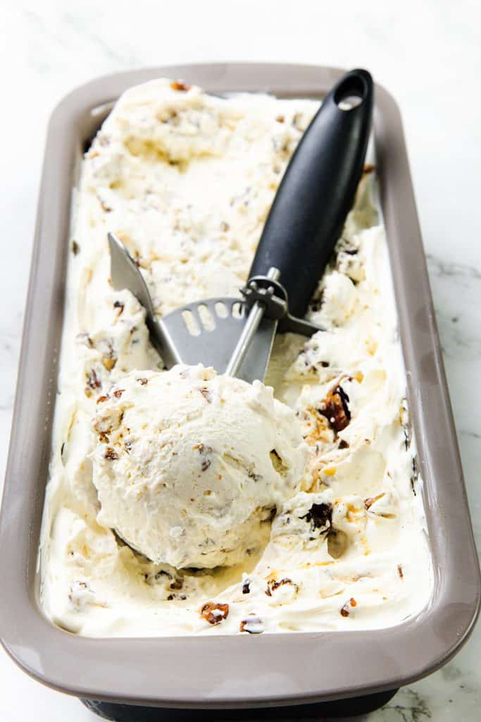 Rectangular container of chunky vanilla ice cream with visible chocolate pieces and hints reminiscent of Butter Pecan Ice Cream, a scoop of ice cream in a metal ice cream scooper rests on top, all on a marble surface.