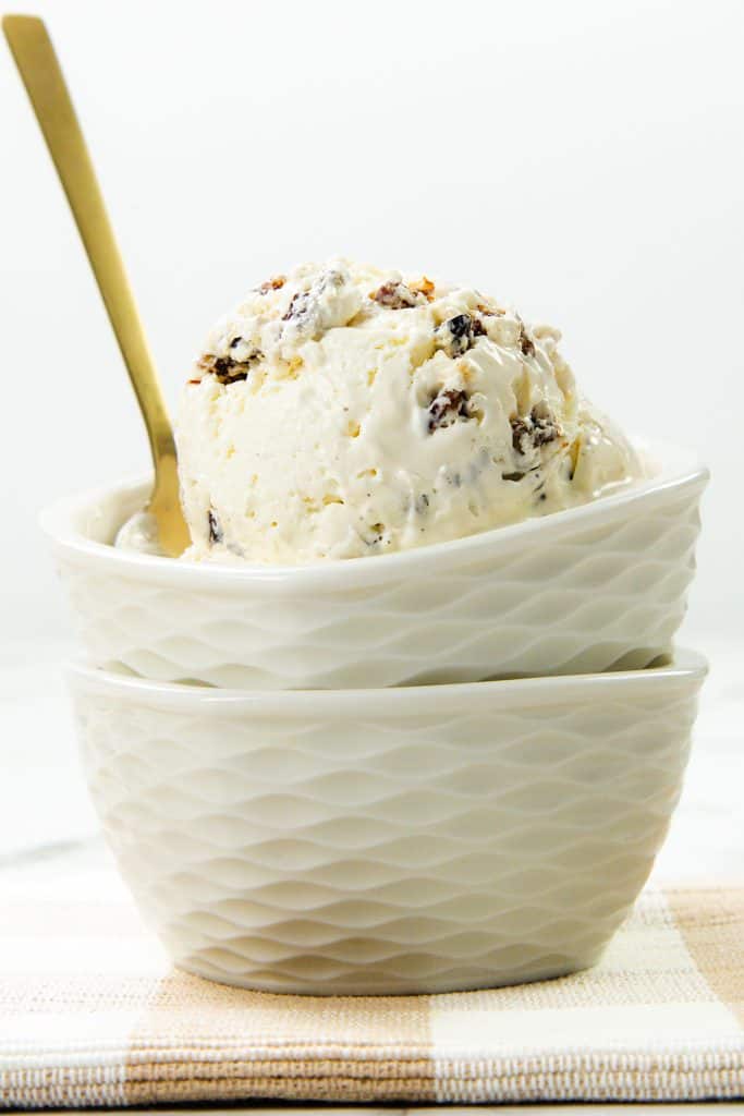 A scoop of rum raisin ice cream sits in a white textured bowl, with a gold spoon, stacked on another empty bowl atop a checkered cloth—the perfect classic pairing for anyone who loves rich flavors like Butter Pecan Ice Cream.