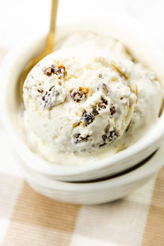 A close-up of a bowl of rum raisin ice cream with a spoon, showing creamy vanilla ice cream mixed with raisins, sits beside a scoop of rich butter pecan ice cream for an irresistible dessert duo.