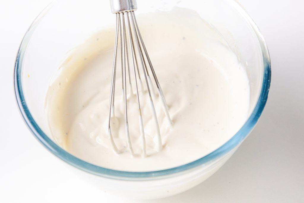 A metal whisk rests in a glass bowl filled with creamy white Caramelized Onion Dip, against a white background.