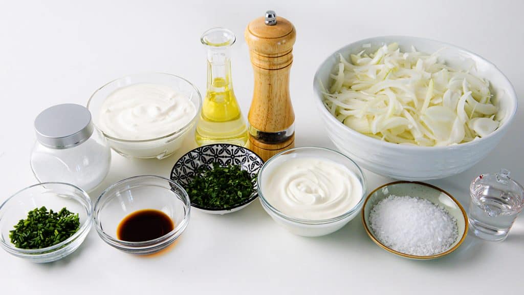 Various ingredients on a white table, including sliced onions in a bowl, sour cream, yogurt, oil, salt, pepper, chopped herbs, soy sauce, and a small dish of water&mdash;perfect for creating a rich and flavorful Caramelized Onion Dip.