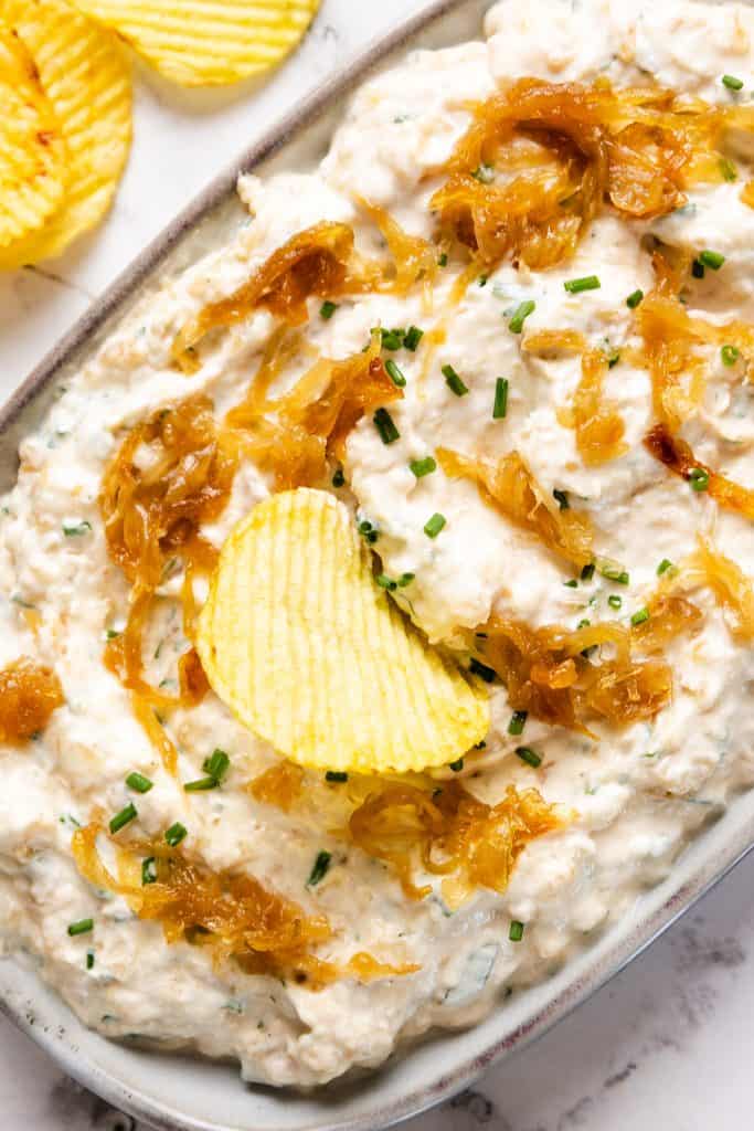 A bowl of creamy Caramelized Onion Dip is topped with sweet caramelized onions and chopped chives, garnished with a ridged potato chip. More chips are placed nearby on the marble surface.