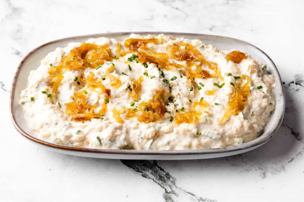 A white oval dish filled with creamy Caramelized Onion Dip, garnished with sweet caramelized onions and chopped chives, sits on a marble surface.