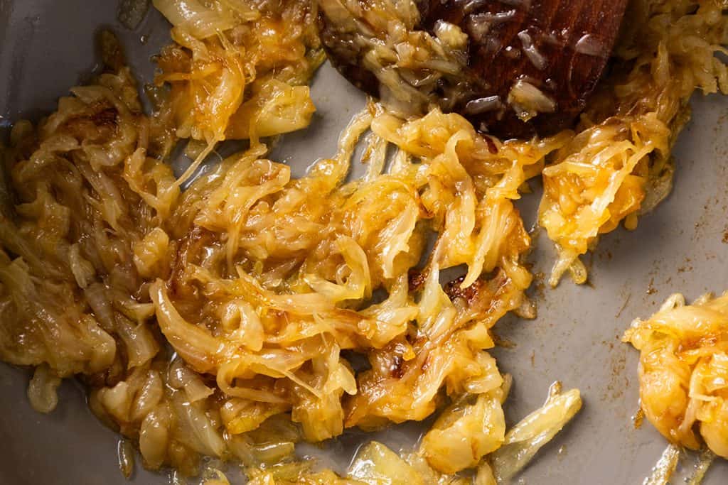 Golden brown caramelized onions cooking in a pan, with a wooden spatula stirring them. The onions appear soft, glossy, and slightly sticky from the caramelization process&mdash;perfect for making a rich and flavorful Caramelized Onion Dip.