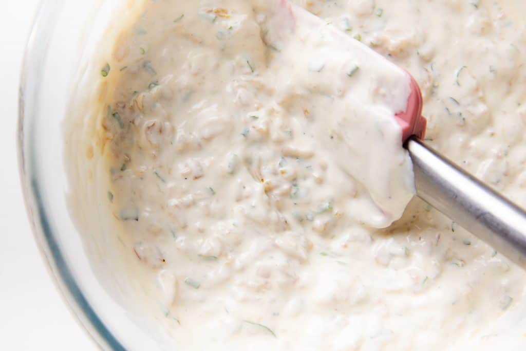 A close-up of a glass bowl filled with a creamy Caramelized Onion Dip containing herbs and spices. A red and white spatula is partially submerged in the mixture.