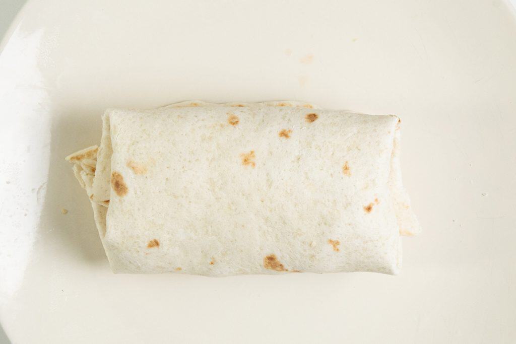 A Chicken Caesar Wrap, snugly rolled in a flour tortilla, sits on a plain white plate viewed from above. The wrap is closed on both ends, and the plate is mostly empty except for a few small crumbs.