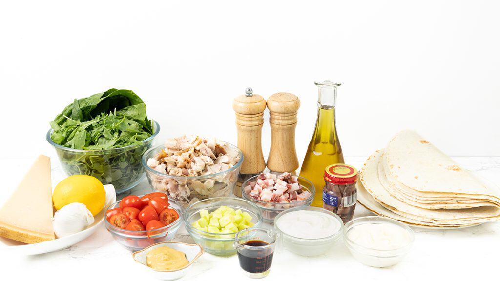 Various fresh ingredients displayed on a white surface, including leafy greens, cherry tomatoes, cheese, cooked chicken for a Chicken Caesar Wrap, diced bacon, lemon, tortillas, seasonings, oil, and sauces in glass bowls.