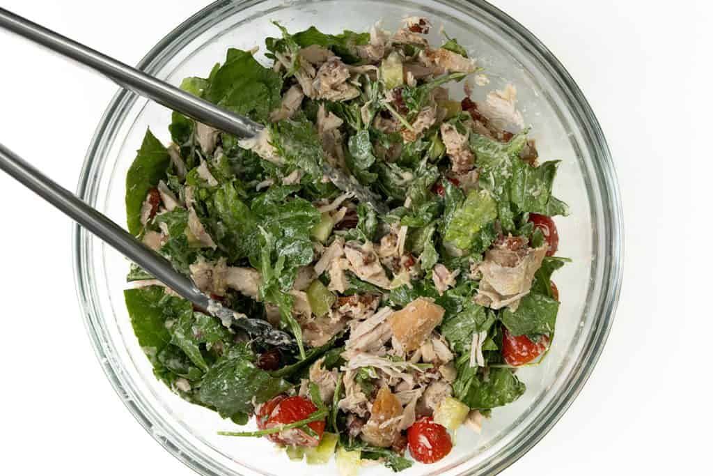 A glass bowl filled with salad featuring shredded chicken, spinach, cherry tomatoes, croutons, and dressing—perfect for enjoying on its own or using as a filling for a delicious Chicken Caesar Wrap. Metal tongs rest inside for easy serving.