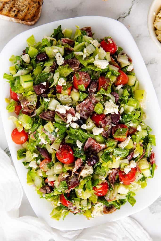 A white plate holds a refreshing chopped salad with lettuce, cherry tomatoes, cucumbers, olives, green peppers, red onions, and crumbled feta cheese.