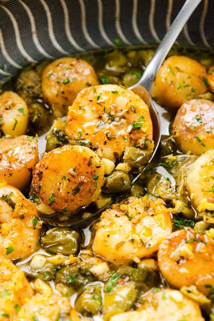Seared scallops, garnished with herbs and served in a flavorful brown butter garlic sauce with capers. A spoon is lifting one scallop from a patterned bowl, highlighting the rich glaze. Perfect alongside pan seared shrimp for an elegant seafood experience.