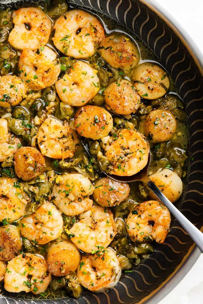 Shrimp and scallops, pan seared to perfection, are cooked in a rich brown butter garlic sauce and garnished with chopped parsley, served in a round dish with a spoon.