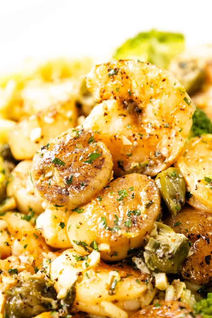 Close-up of pan seared shrimp garnished with herbs and spices, mixed with chopped vegetables including broccoli and capers, all drizzled with a rich brown butter garlic sauce.
