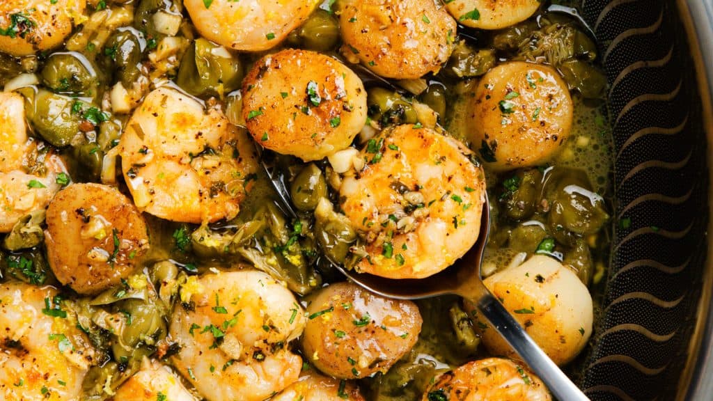 Pan-seared scallops in a pan with brown butter garlic sauce, green herbs, and chopped vegetables, with a spoon partially visible on the right side.