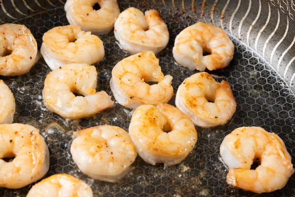 Pan seared shrimp are being sautéed in a frying pan, lightly browned and seasoned, with bubbles indicating they are being cooked over heat.