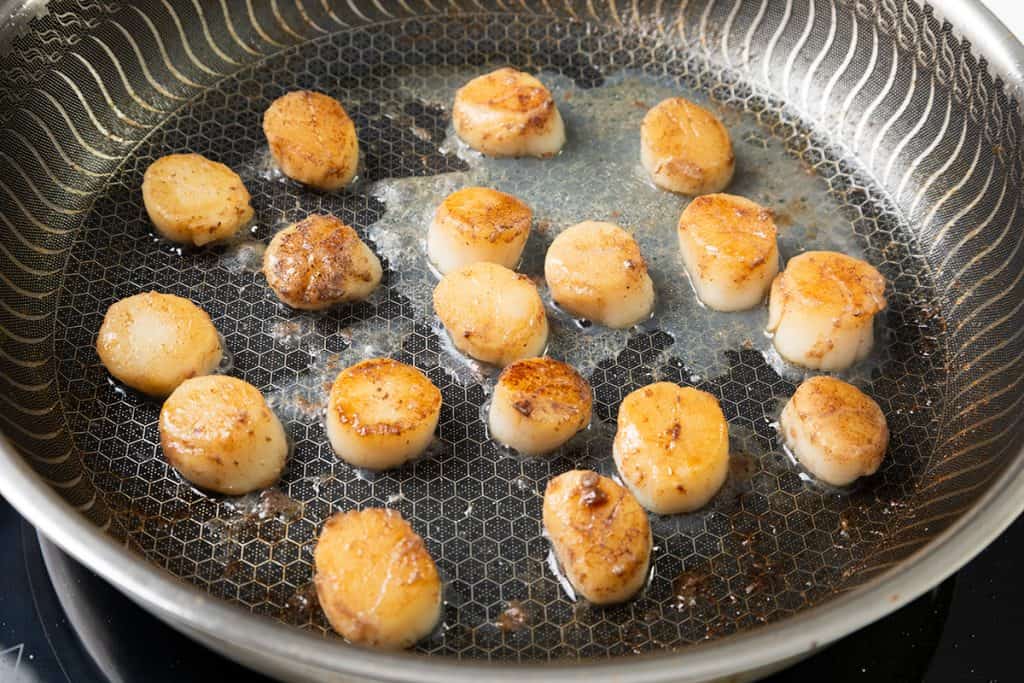Several scallops are being seared in a stainless steel pan, showing a golden-brown crust on their surfaces. The pan is on a stovetop, and some oil is visible around the scallops—perfect for preparing an Easy Shrimp and Scallops recipe.