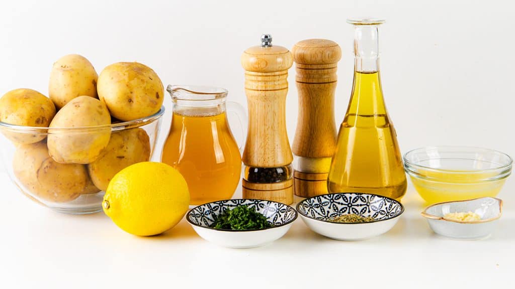 A bowl of potatoes, a lemon, oil, vinegar, chopped herbs, salt, pepper grinders, minced garlic, and a bowl of yellow liquid are arranged on a white surface—perfect ingredients for preparing classic Greek Lemon Potatoes.