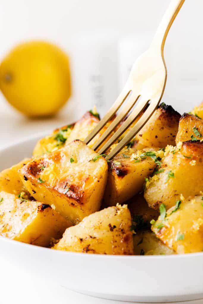 A fork spears a roasted potato in a bowl filled with golden, seasoned Greek Lemon Potatoes, garnished with chopped herbs. A lemon and salt shaker are blurred in the background.