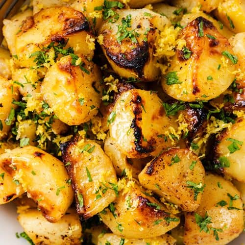 Greek Lemon Potatoes: Golden roasted potatoes garnished with chopped parsley and lemon zest, served in a white bowl with a gold fork on the side.