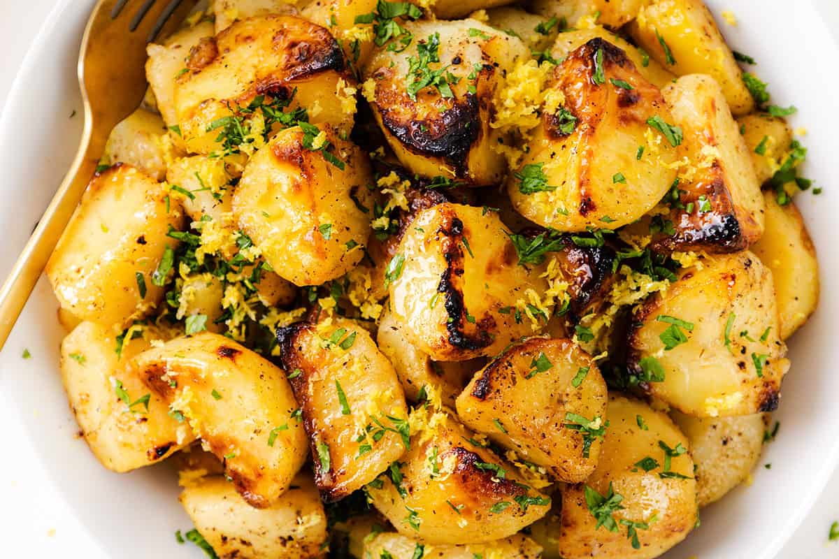 Greek Lemon Potatoes: Golden roasted potatoes garnished with chopped parsley and lemon zest, served in a white bowl with a gold fork on the side.