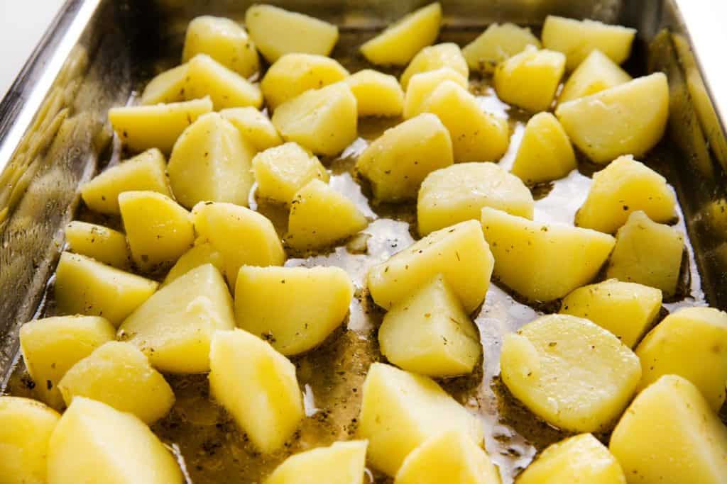 Chopped potatoes seasoned with herbs, oil, and a burst of zesty lemon are spread out in a metal baking dish, ready to be roasted into classic Greek Lemon Potatoes.